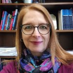 Individual with long hair and colorful scarf sits before wooden bookshelf filled with books, documents, and a small plant on the top shelf
