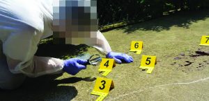 Forensic examiner in protective suit and blue gloves inspects ground evidence with magnifier beside yellow numbered markers 3–6, rope line, and outdoor surface