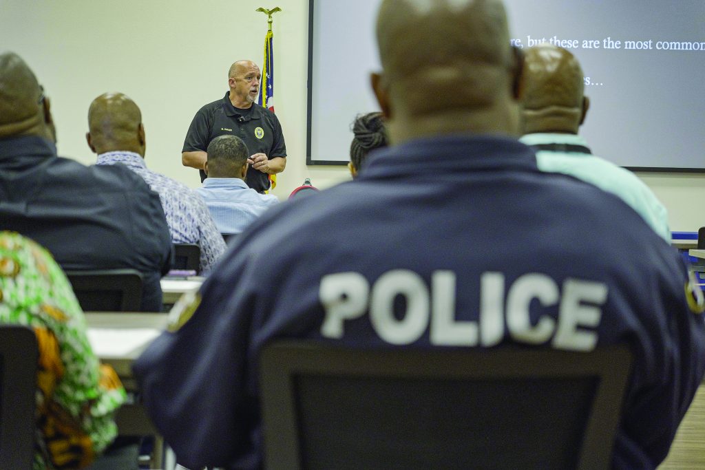 a classroom setting with several individuals seated and facing forward, listening to a speaker at the front of the room. The speaker, who is standing near a podium, is wearing a black shirt with an emblem on it. The faces of the individuals are not clearly visible as they are mostly turned away from the camera. One individual in the foreground has "POLICE" written in large white letters on the back of their dark blue shirt. There is an American flag positioned behind the speaker, and part of a projection screen displaying text can be seen on the right side of the image.