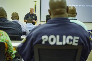 a classroom setting with several individuals seated and facing forward, listening to a speaker at the front of the room. The speaker, who is standing near a podium, is wearing a black shirt with an emblem on it. The faces of the individuals are not clearly visible as they are mostly turned away from the camera. One individual in the foreground has "POLICE" written in large white letters on the back of their dark blue shirt. There is an American flag positioned behind the speaker, and part of a projection screen displaying text can be seen on the right side of the image.