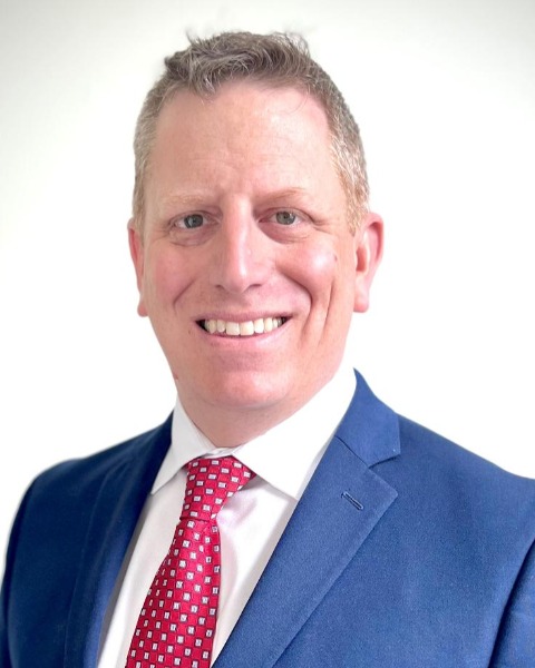 Individual in blue suit jacket, white dress shirt, and red patterned tie posed against plain white background