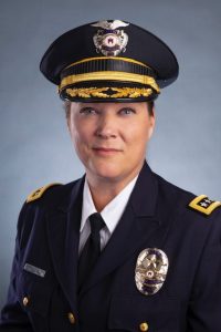 Police officer in dark formal uniform with gold trim, tie, badge, and peaked cap featuring emblem, standing against a plain gray background