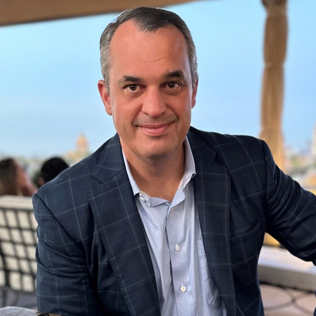Man in dark checkered blazer and light blue shirt sits under canopy with beige curtains; outdoor setting with blurred buildings and people behind.