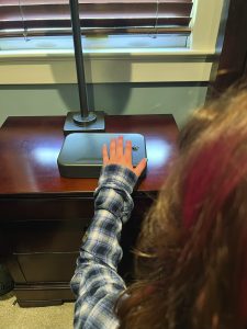 Person in plaid shirt reaching toward a small gray lockbox on a wooden nightstand with lamp, near window with closed blinds in a home setting