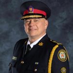 Toronto Police officer in formal uniform with gold epaulettes, left-arm badge, and name tag; posed against textured dark grey background
