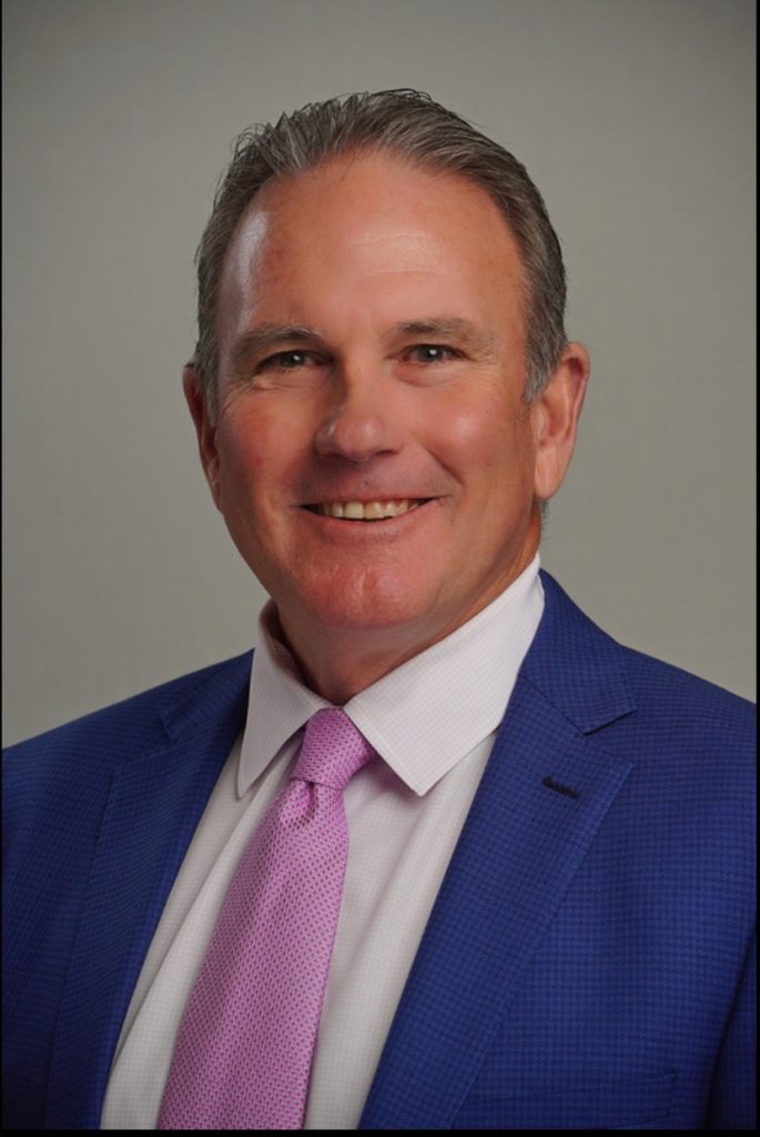 A professional headshot of a person. The individual is wearing a blue suit jacket, a white dress shirt, and a pink tie. The background is plain and gray, providing no additional context or details. The person's hair appears to be short and gray.