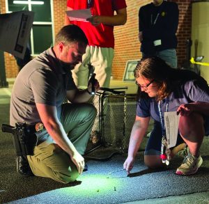 Two individuals examine ground evidence under flashlight; one holds clipboard, other uses tweezers; others stand nearby against brick wall.
