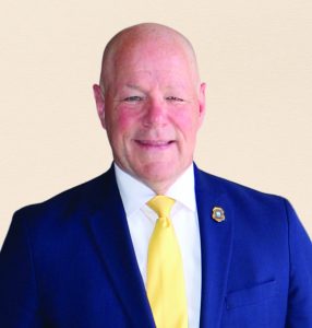 Person wearing a dark blue suit jacket with a white dress shirt and bright yellow tie, featuring a small circular lapel pin on the left side