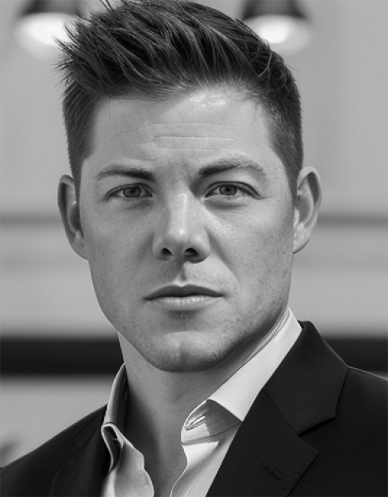 Black‑and‑white portrait shows short styled hair, suit jacket, open‑collar shirt, and clean background, with sharp focus, shallow depth, and studio lighting