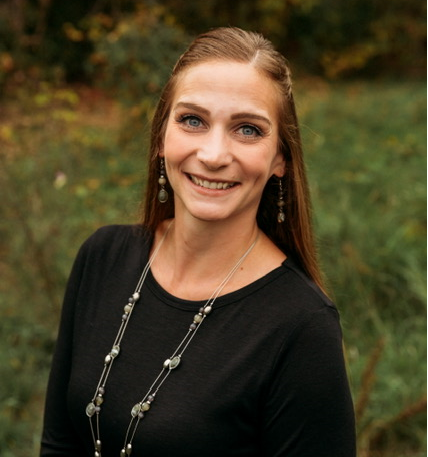 Person wearing a long-sleeve black top and layered bead necklace stands outdoors in front of green grass and autumn foliage in soft natural light.