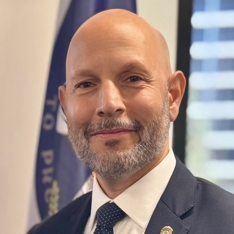 Individual in a dark suit jacket, white dress shirt, and dotted tie with shield-shaped lapel pin stands indoors before a flag, window blinds, and office setting with soft natural light