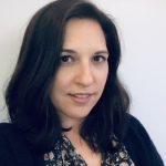 Person wearing a dark cardigan over a floral-patterned blouse with pink and green accents, standing against a plain light-colored wall background.