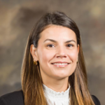 Individual with long brown hair in black blazer and white lace-trimmed blouse stands before a softly blurred brown and beige-toned background.