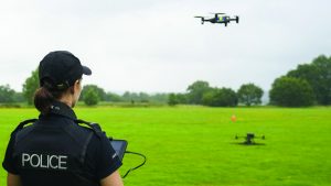 Uniformed police operator controls a quadcopter drone using a tablet in an open grassy field, with a second drone on the ground, trees, cones, and cloudy sky visible