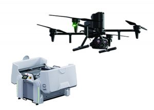 Black quadcopter drone with mounted multi-lens camera and green indicator light shown above a white automated docking station with control panel and open bay.