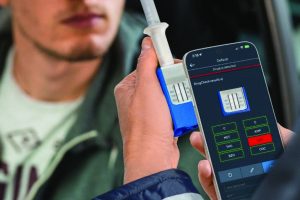 Hands hold a DrugCheck saliva test device and smartphone displaying drug screening results, with labeled substance categories, test strip image, vehicle interior, and seated subject visible in background