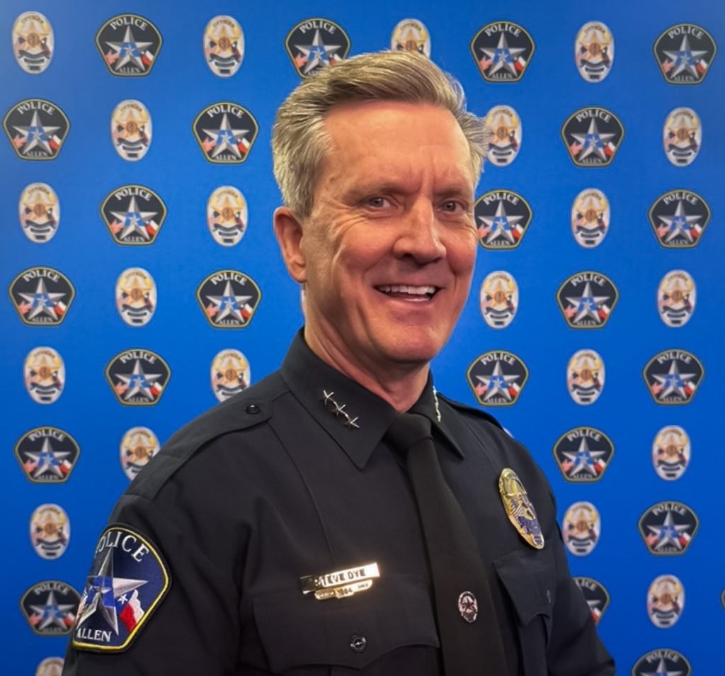 Person wearing a black police uniform with silver collar pins, nameplate, and badge stands before a blue backdrop patterned with police star emblems