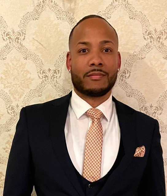 Person wearing a dark suit with white dress shirt, patterned peach tie, and matching pocket square stands against a beige wall with ornate design