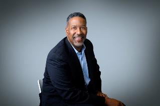Individual in dark suit and light blue shirt seated on white chair against gradient gray background