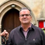 Person wearing a black jacket over a checkered maroon shirt, holding a black metal object, standing in front of an arched stone doorway with a red sign