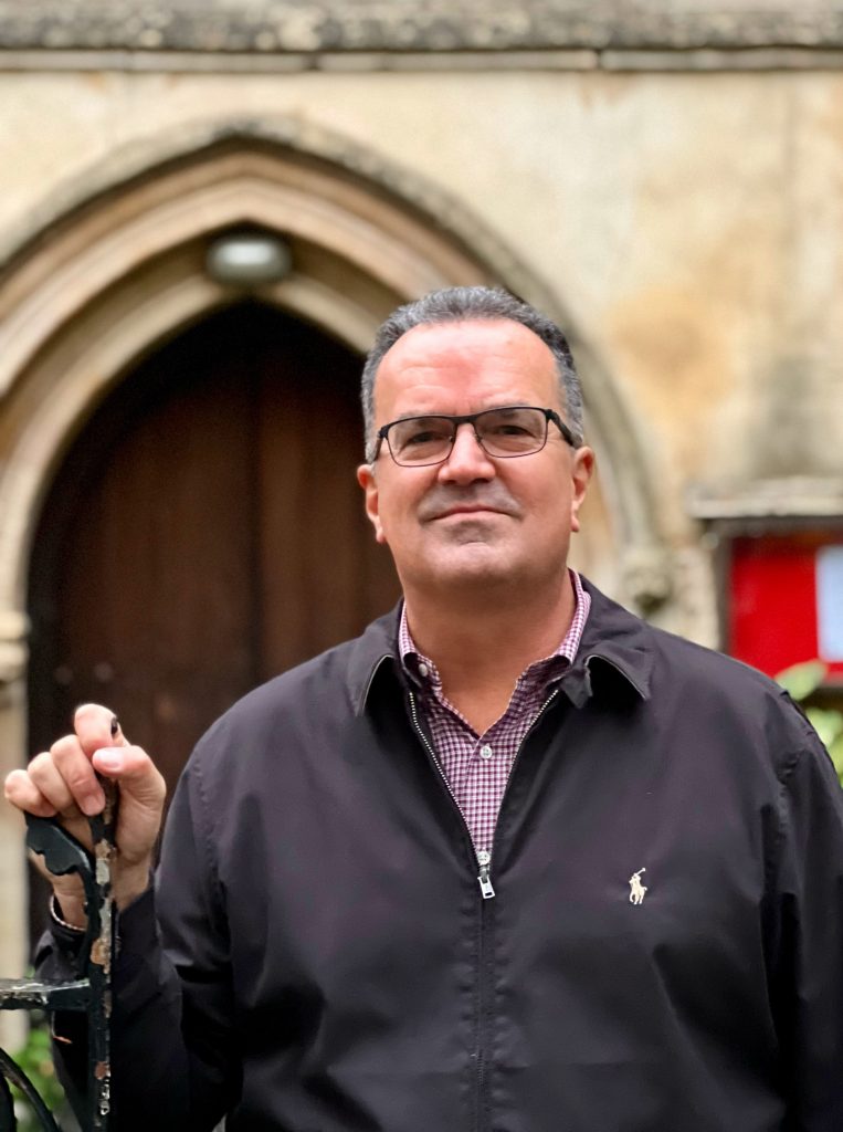 Person wearing a black jacket over a checkered maroon shirt, holding a black metal object, standing in front of an arched stone doorway with a red sign