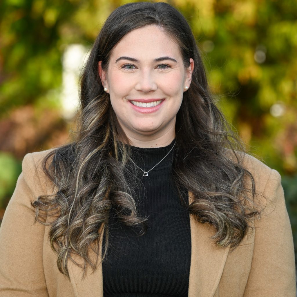 a person with long, wavy hair standing outdoors in front of a blurred background of green foliage. The person is wearing a black turtleneck sweater and a tan coat. A delicate necklace with a small pendant is visible around the person's neck.