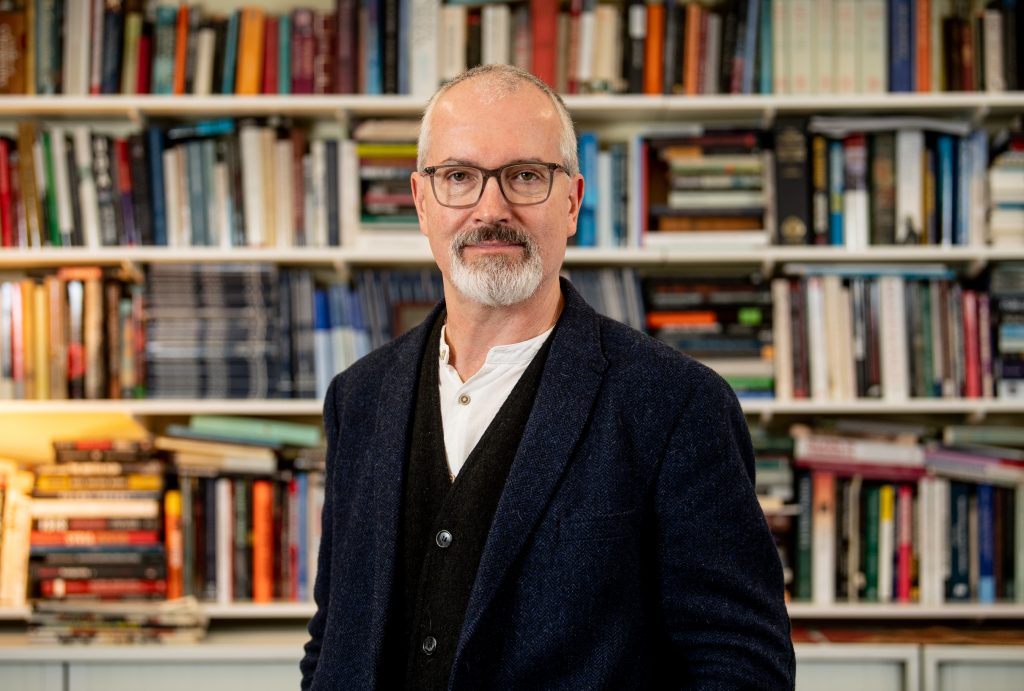 An individual wearing a dark jacket over a buttoned shirt stands in front of shelves filled with books of various sizes and colors in a well‑lit indoor setting