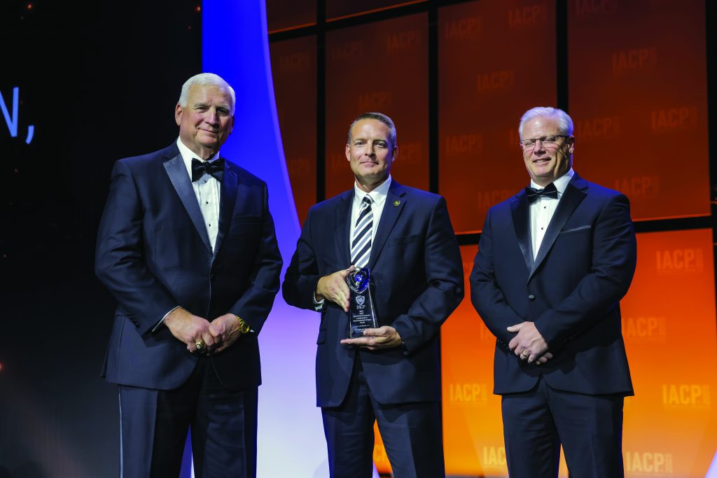 Three individuals in formal black tuxedos stand on stage with orange backdrop and IACP logo; center person holds a clear glass award under bright lighting.