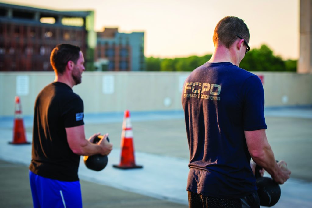 two individuals exercising outdoors on a rooftop or parking structure. Both are holding kettlebells and appear to be engaged in a workout. The individual on the left is wearing a black shirt and blue shorts, while the individual on the right is wearing a navy blue shirt with "FCPD" written on the back, along with black shorts. There are three orange traffic cones placed in the background, and an urban landscape with buildings can be seen beyond the edge of the rooftop. The lighting suggests it is either early morning or late afternoon. The faces of both individuals are visible, showing focused expressions as they perform their exercises.