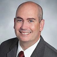 Person in a gray suit jacket with a white shirt, red tie, and a small law‑enforcement‑style lapel pin posed against a neutral gray studio background.