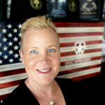 A person with short blonde hair is standing in front of an American flag backdrop. Behind the person, there are several books displayed on a shelf. The visible book titles include "RELENTLESS COURAGE" and "WARRIOR." There is also a logo on the flag backdrop that resembles a shield with a cross and the text "BOOK BANNED."
