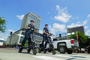 Two uniformed officers riding black electric patrol scooters in a city crosswalk, with tall office buildings, traffic lights, and a white pickup truck nearby