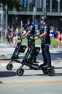 Two uniformed police officers wearing helmets patrol on black electric scooters marked ‘Fargo Police’ near fountains in a busy outdoor public area