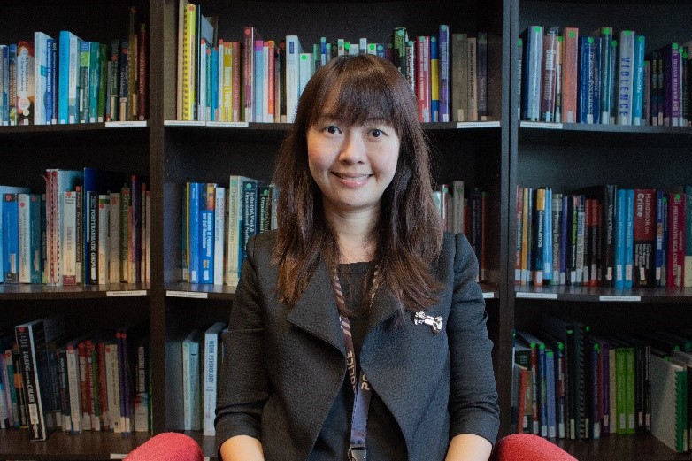 Individual wearing a dark blazer with decorative brooch and conference lanyard sits before tall bookshelves filled with books, framed by red chairs and library furnishings