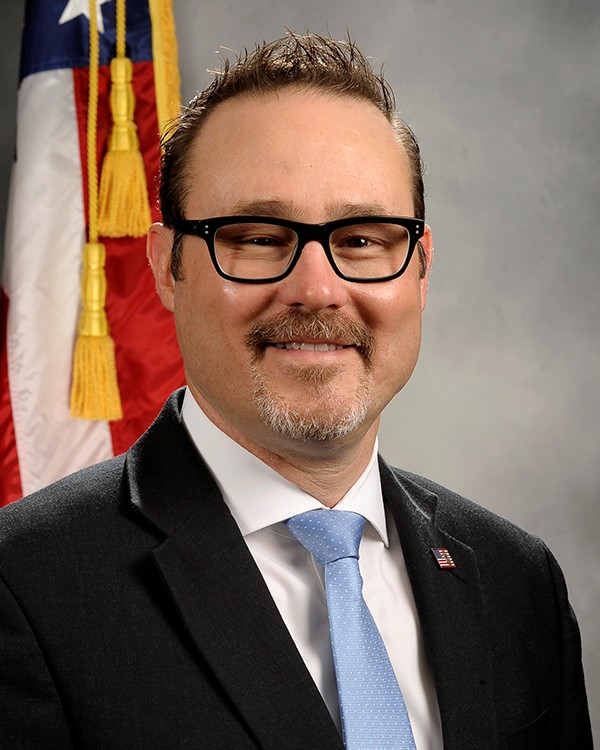 Individual wearing black suit jacket, white shirt, light blue tie, rectangular eyeglasses, and flag lapel pin stands before U.S. flag and gray studio backdrop