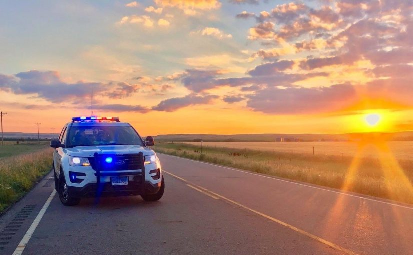 Marked police SUV with flashing red and blue emergency lights is parked on a rural two-lane roadway at sunset, bordered by grasslands, utility poles, fences, rolling hills, and a colorful sky with clouds and sun rays stretching across the horizon