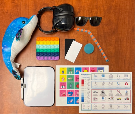 Table with blue dolphin plush, black headphones, sunglasses, rainbow pop-it, whiteboard, visual cards, and assorted sensory communication tools