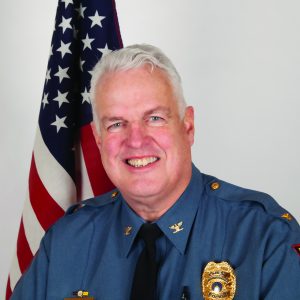 Law enforcement officer in blue uniform with gold insignia and badge stands before U.S. flag; formal portrait-style setting.