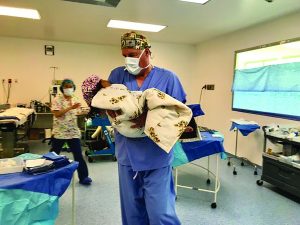 Professional in blue scrubs and surgical cap holds swaddled newborn in OR; another person stands near equipment and surgical tables.