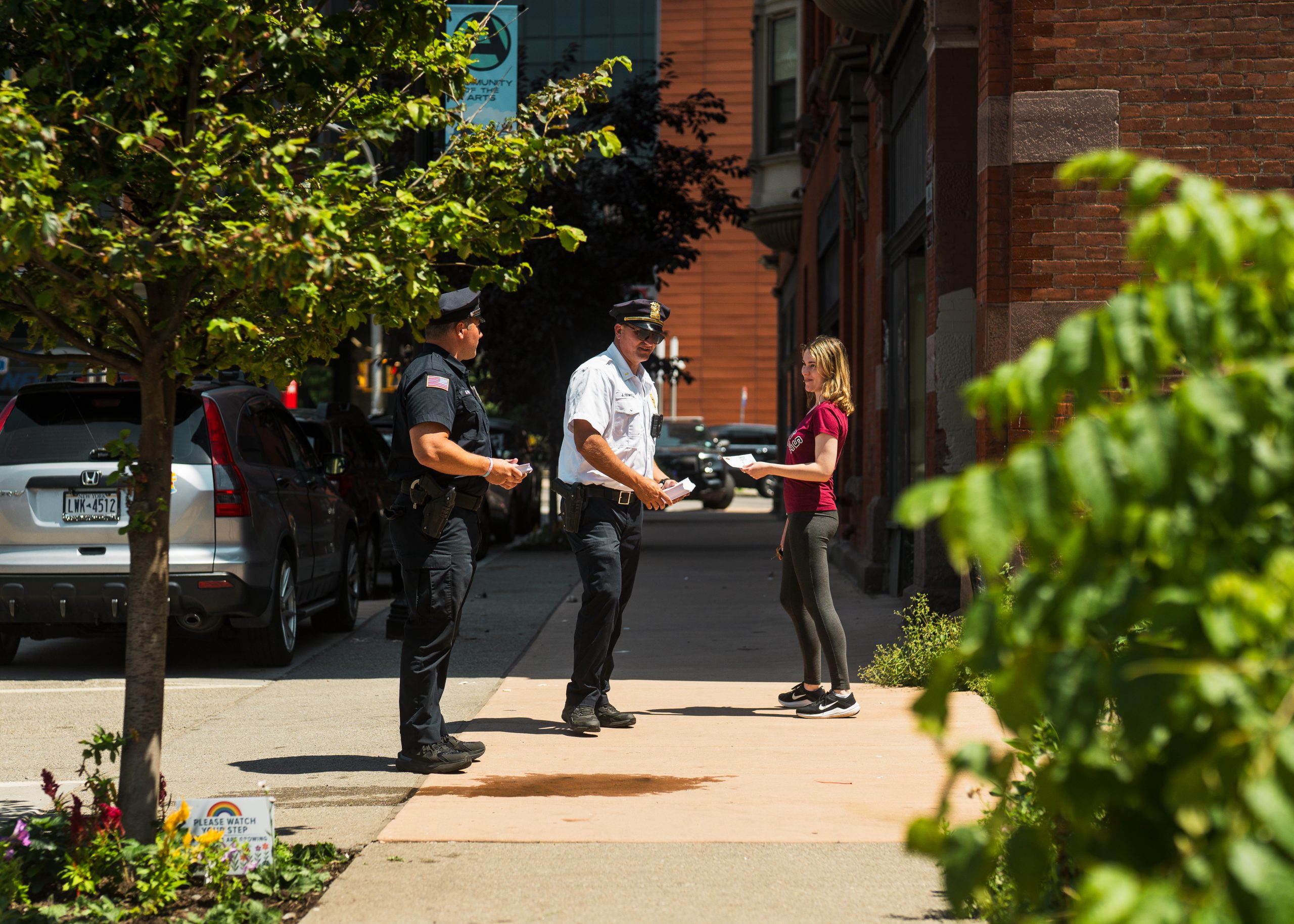 Two uniformed officers stand on urban sidewalk engaging with casually dressed woman; trees, parked cars, and buildings visible in background