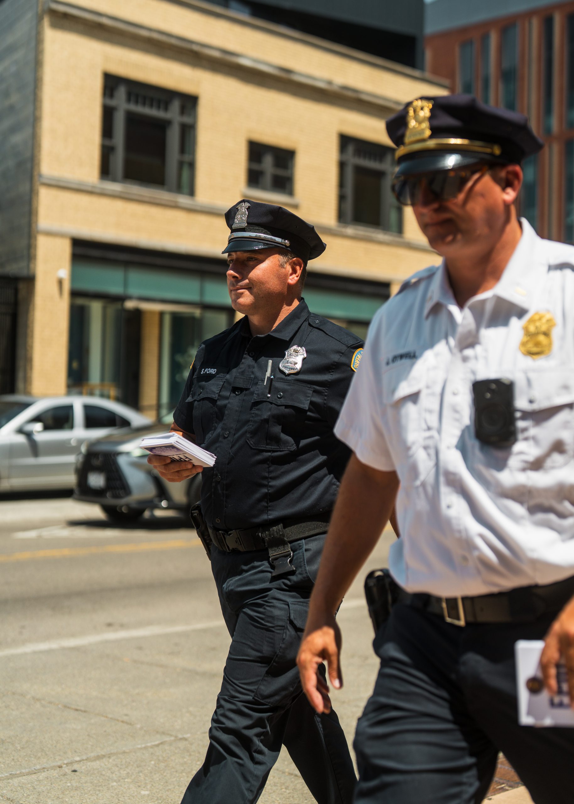 Two uniformed officers walk on city sidewalk, one in black and one in white, holding papers; beige building and parked cars in urban background