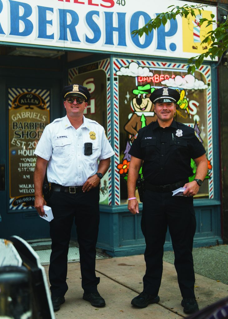 Two uniformed officers stand outside Gabriel's Barbershop, holding papers; storefront features colorful sign and cartoon character on window.