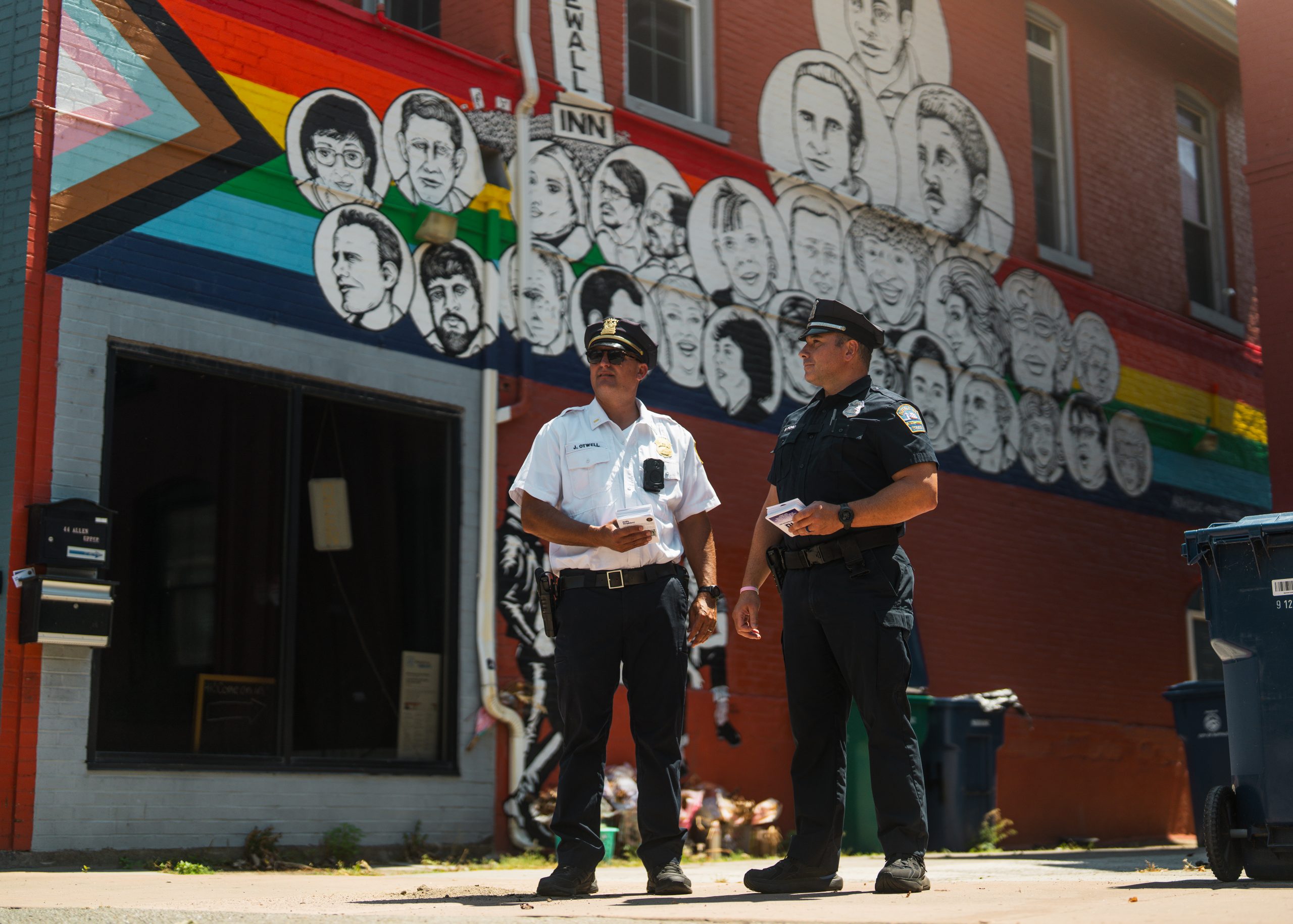 Two uniformed officers stand before brick mural with rainbow flag and illustrated faces, highlighting community engagement and inclusive policing