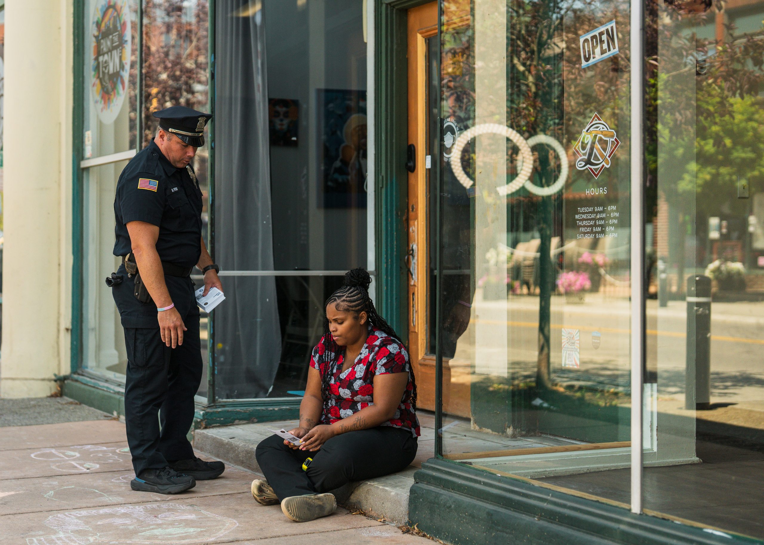 Police officer stands outside glass storefront with 'OPEN' sign, speaking to seated individual on sidewalk; scene includes glass doors and windows.