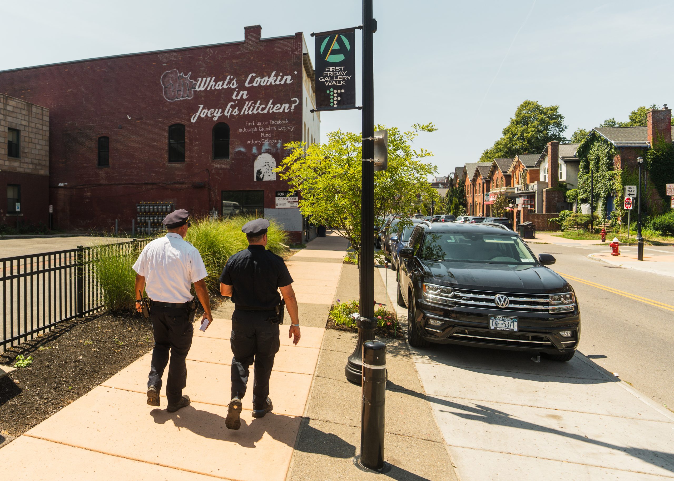 Two uniformed officers walk past parked SUV and mural reading 'What's Cookin' in Joey B's Kitchen?' on brick wall; trees, homes, and art district banner.