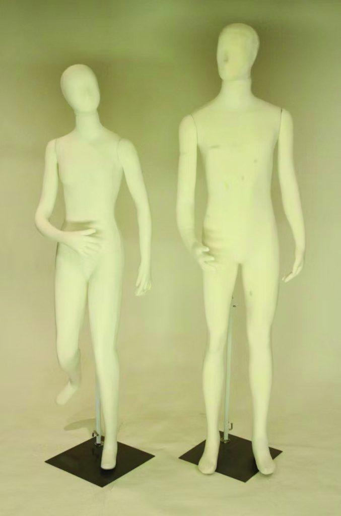 Two white articulated forensic mannequins stand on black metal bases against a neutral backdrop, one posed walking with raised leg and hand at waist, the other upright with arms lowered, showing smooth featureless heads, jointed limbs, torso contours, support rods, and display stands