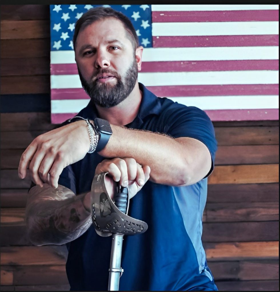 Person in dark polo shirt rests tattooed arms on a ceremonial sword hilt, wearing a watch and bracelets, standing before a wooden wall with a large American flag.