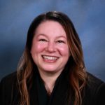 Person with long wavy hair in dark pinstripe suit jacket and dark green top stands against blue gradient background