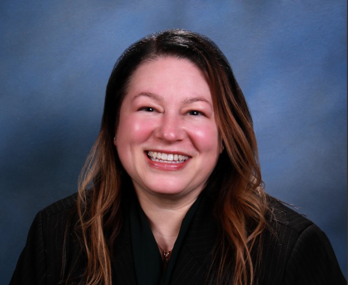 Person with long wavy hair in dark pinstripe suit jacket and dark green top stands against blue gradient background