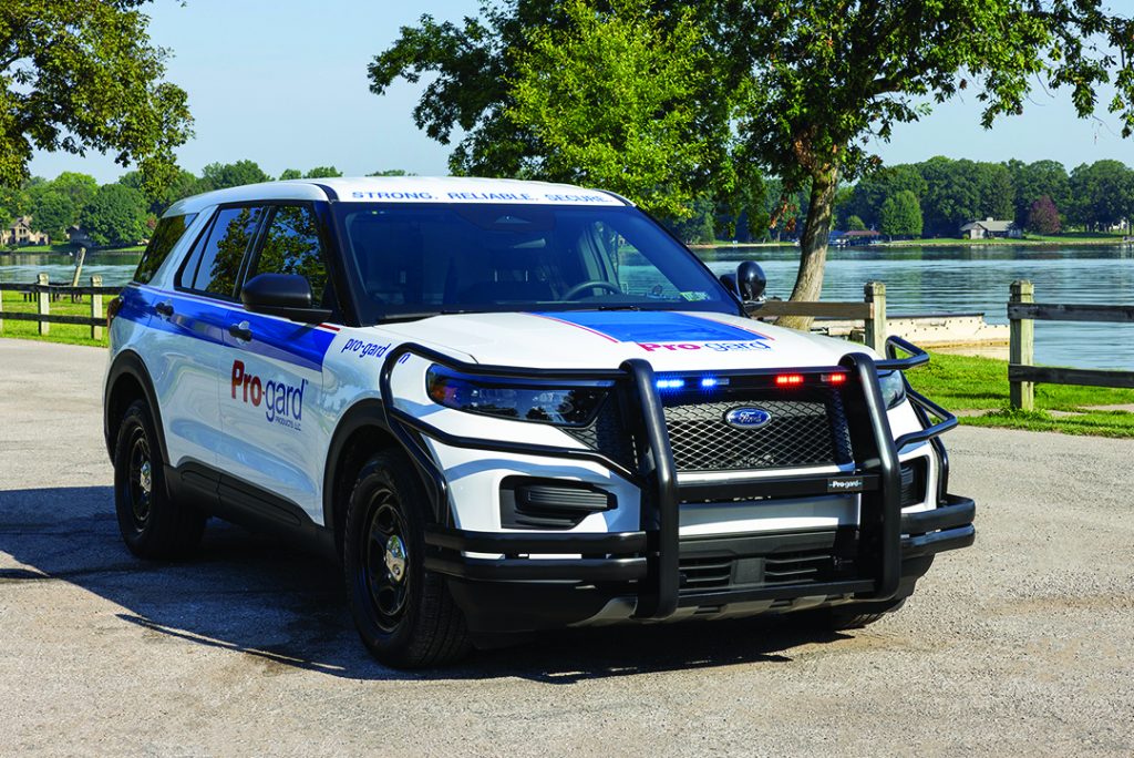 White Ford police SUV with black push bumper, roof lights flashing red and blue, Pro-gard branding, parked near lake with trees and wooden fence.